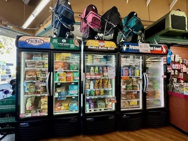 Refrigerated display cases in a pet store, filled with food items. Backpacks and a pet carrier are on top.
