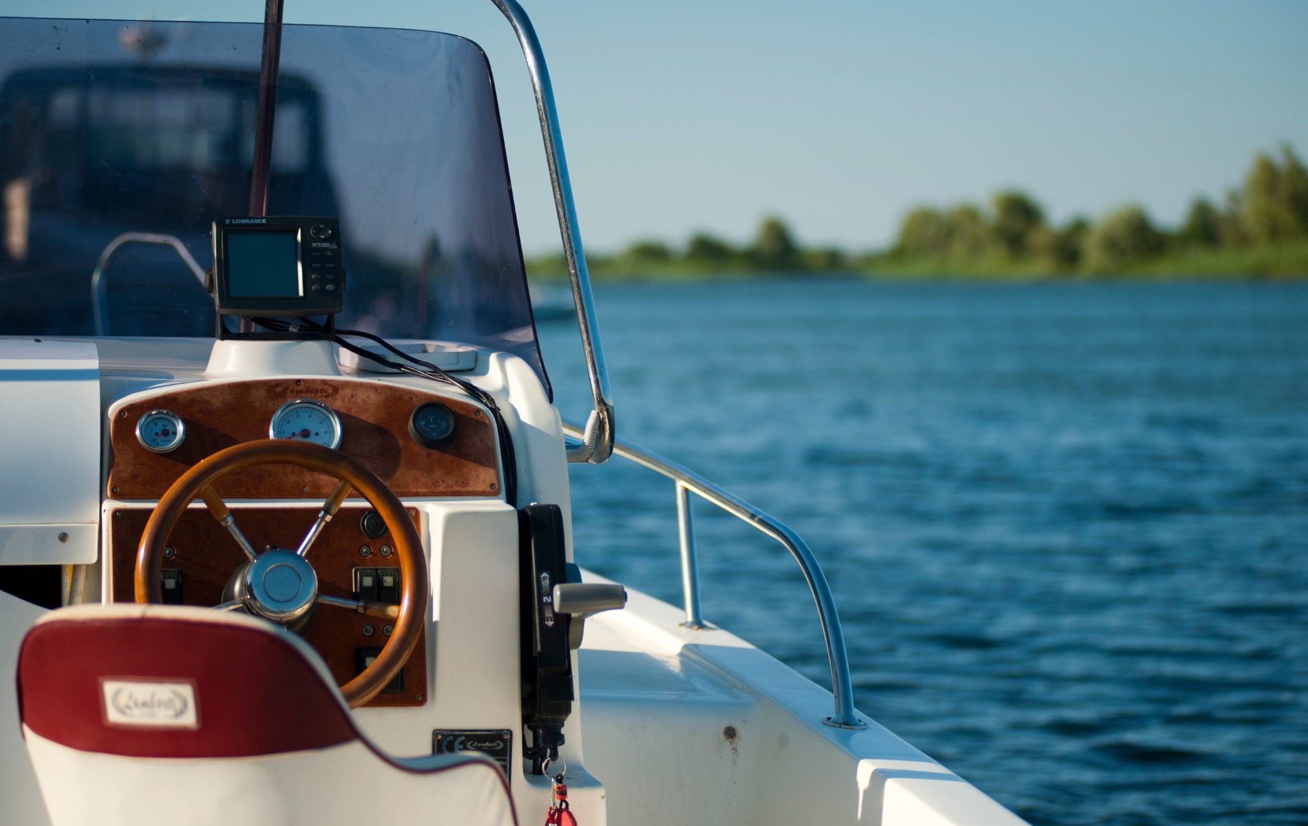 Boat's helm with wooden wheel, dashboard, and seat; blue water and shoreline in the background.