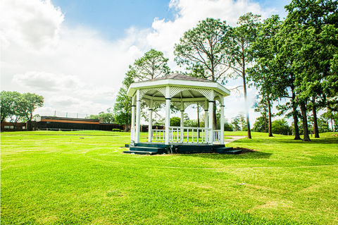 White gazebo in a green grassy park, surrounded by trees under a partly cloudy sky.