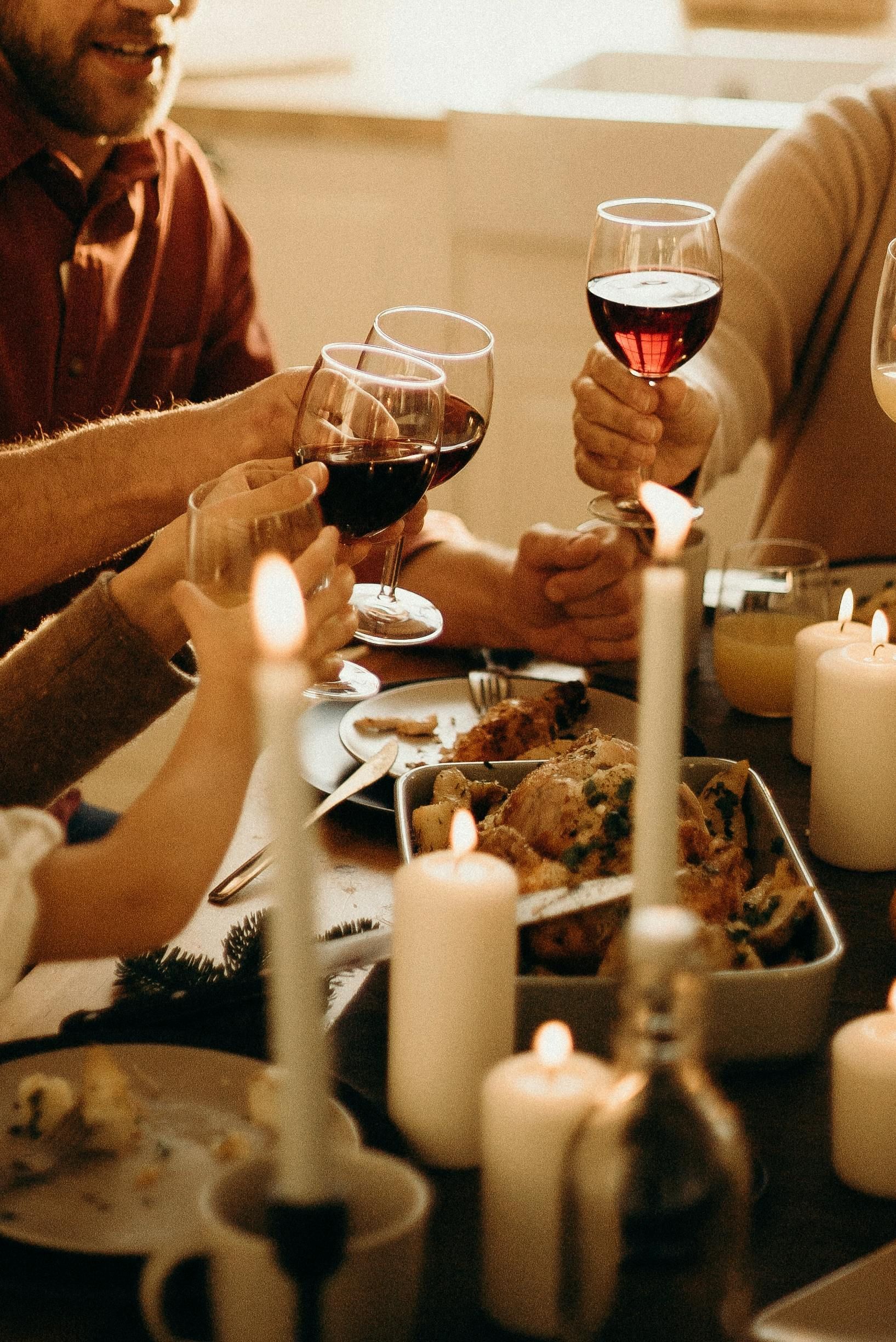 People around a dimly lit table raising wine glasses for a toast over a shared meal, surrounded by lit candles.