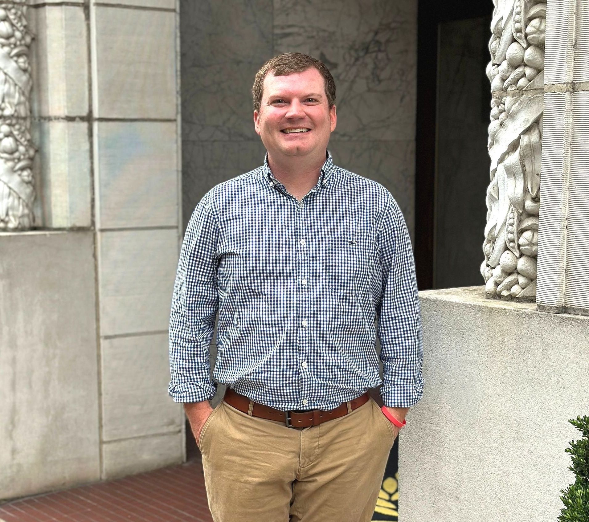 Man with hands in pockets smiles in front of a light gray building. He wears a plaid shirt and khaki pants.