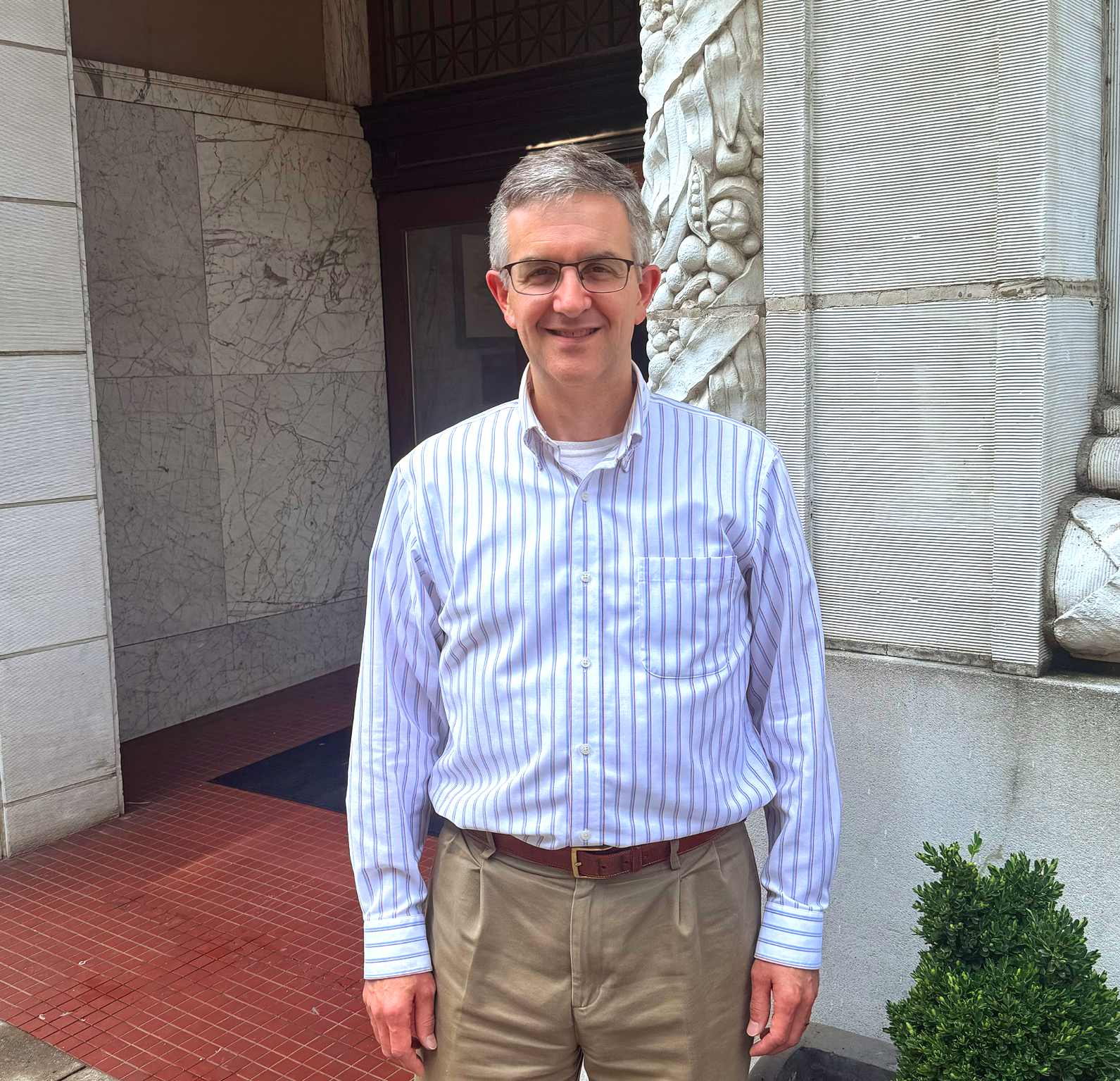 Man in striped shirt and glasses stands outside a building with decorative stone.