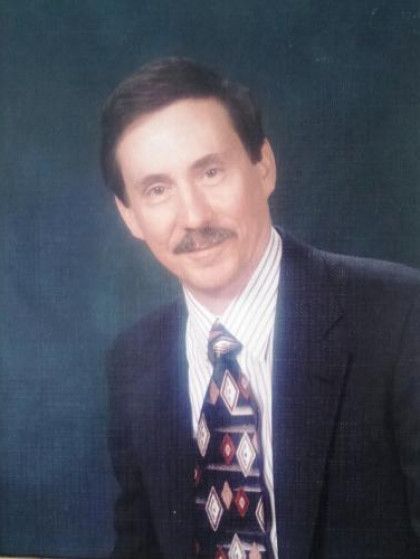 Man in a suit and tie, smiling slightly, with dark hair and a mustache, against a blue background.