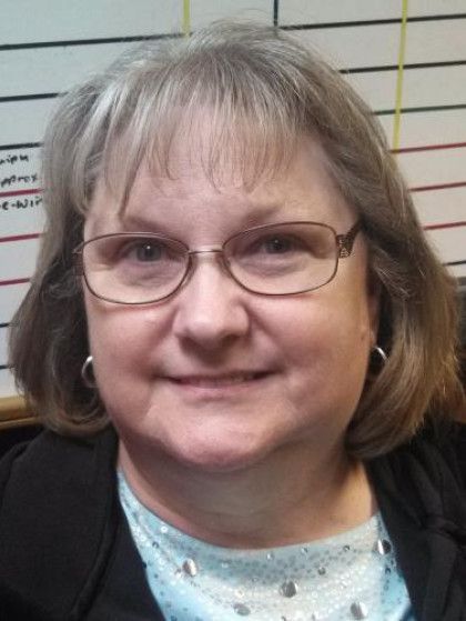 Woman with glasses, smiling, in front of a wall, wearing a black blazer and light blue shirt.