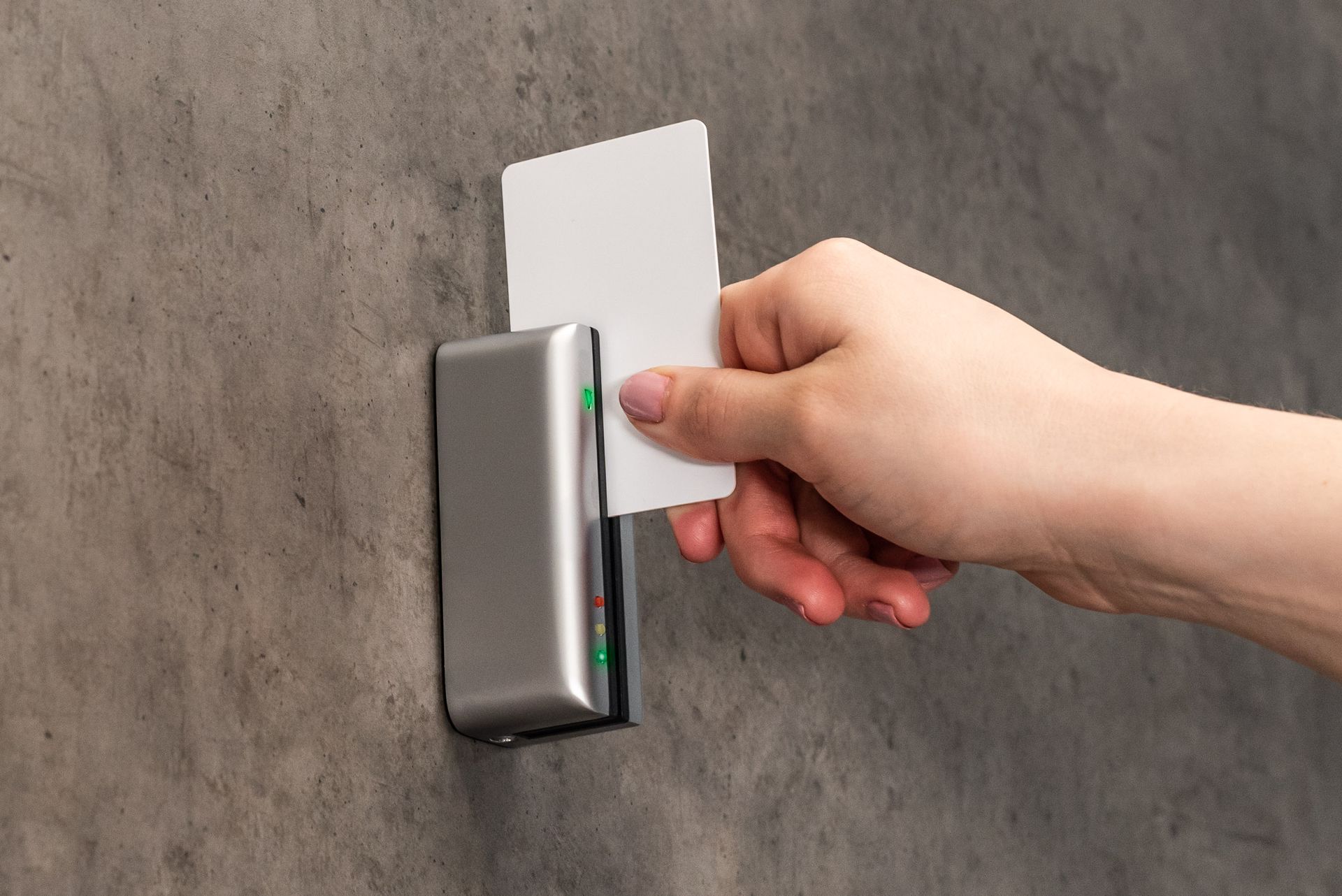 Hand holding a white card near a silver card reader on a gray wall.