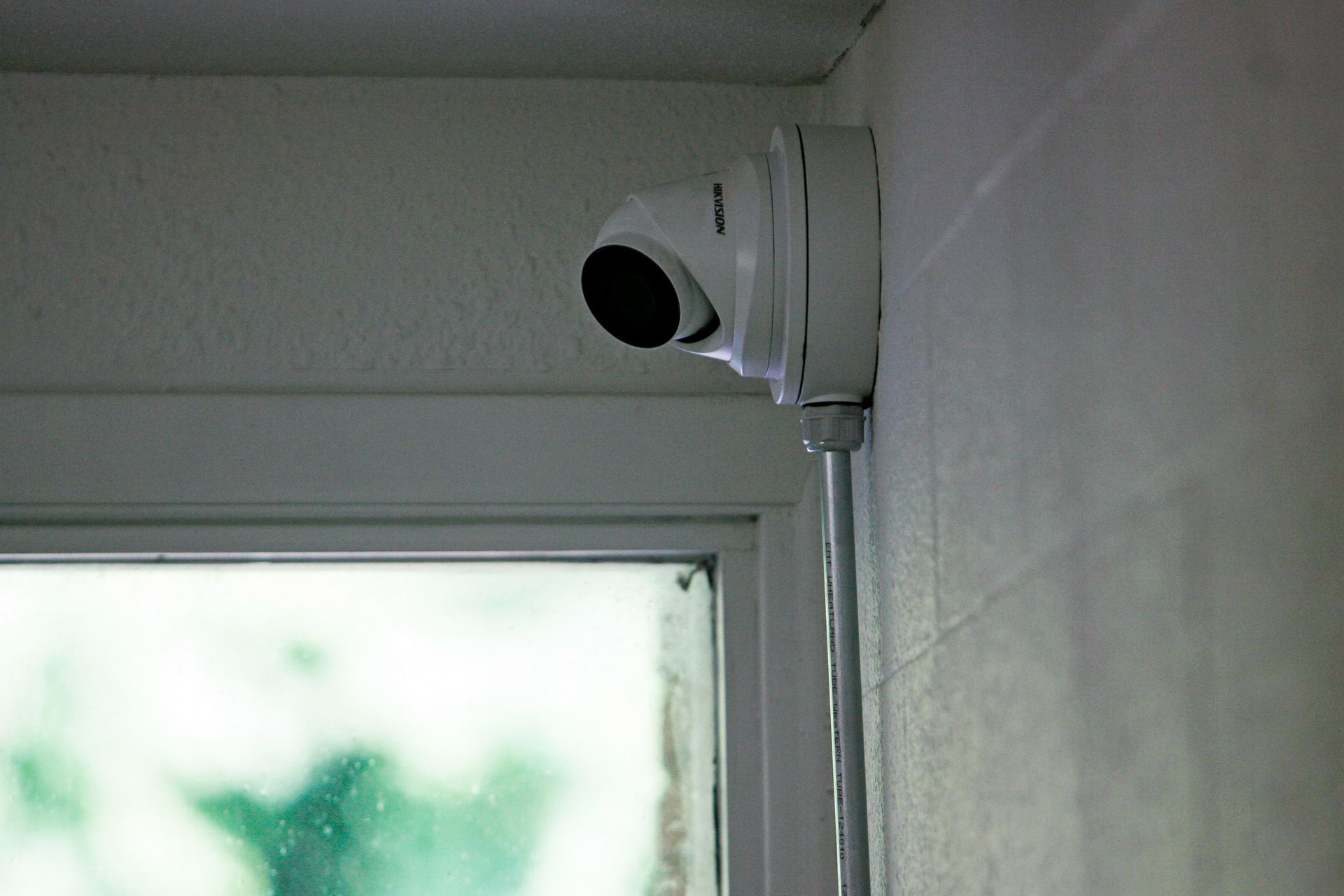 Security camera mounted on a white wall, angled toward a window, with a cable running down.