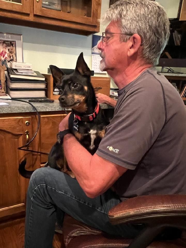 Man in glasses holding small black and brown dog on his lap; indoors, desk in background.