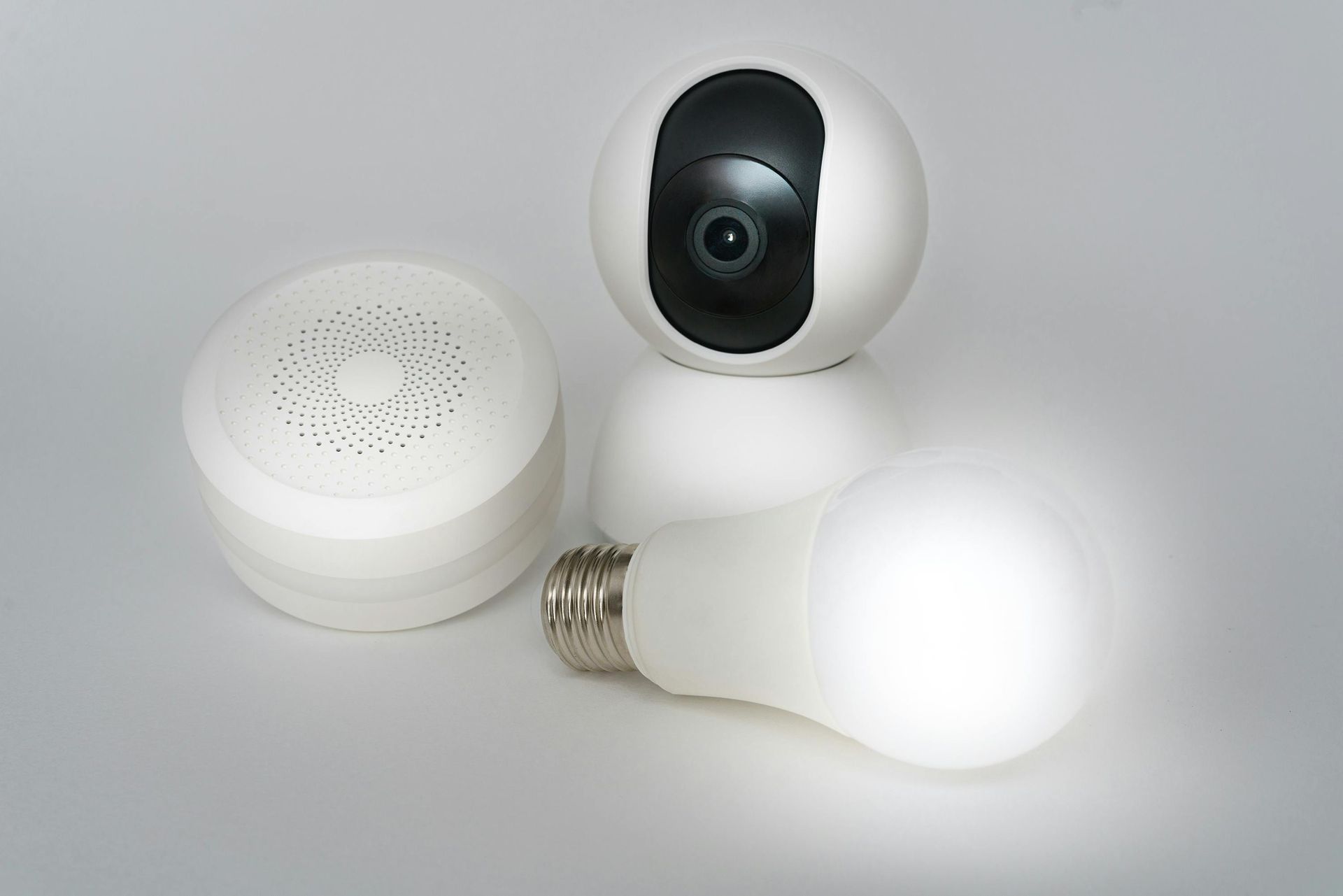 White security camera, smart speaker, and lit LED light bulb on a light gray surface.