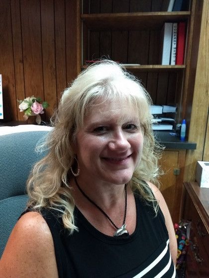 Woman with blonde hair in office setting, smiling. Wearing black sleeveless top, jewelry. Wooden shelves in background.