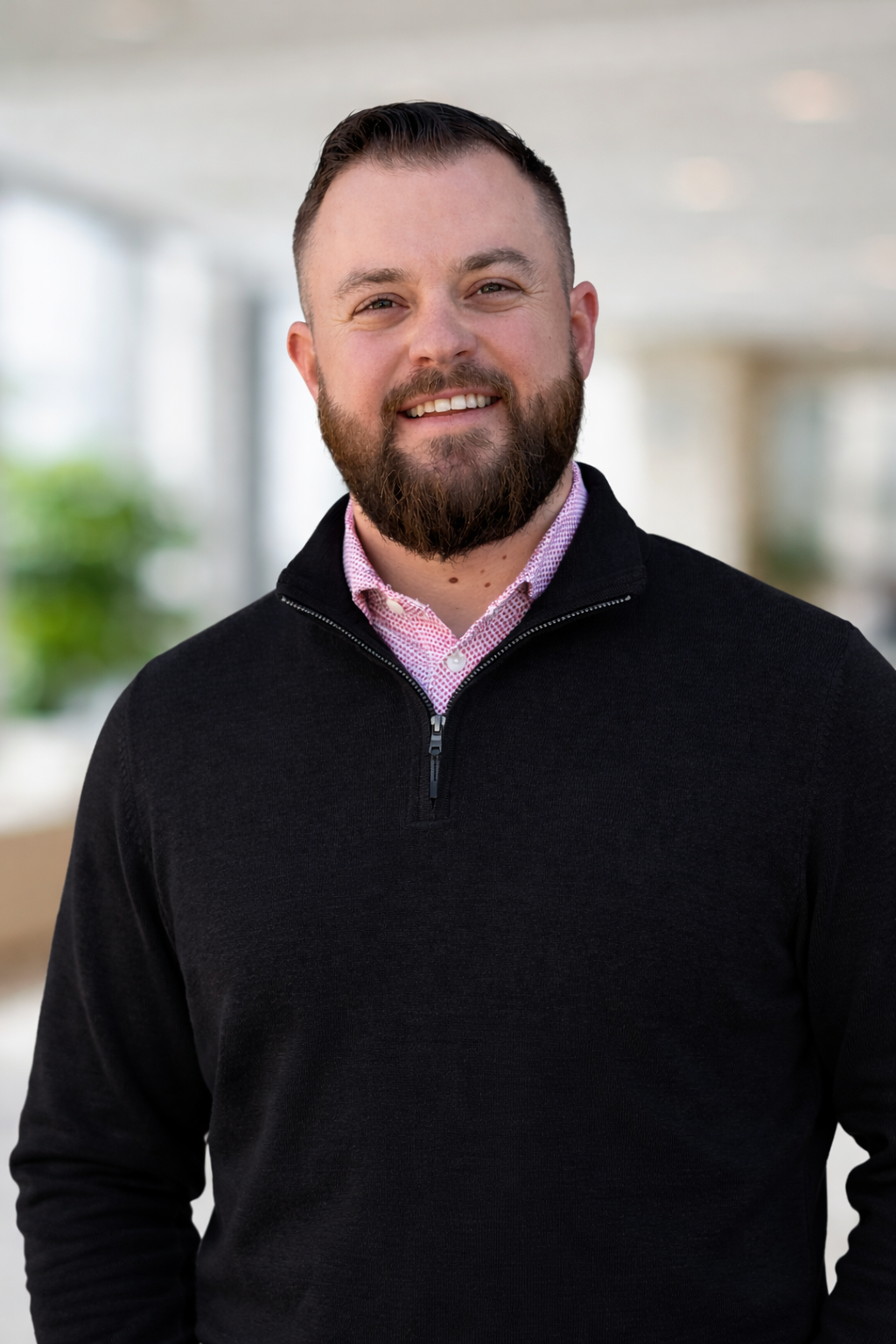 Smiling man in a black sweater over a pink shirt, standing indoors against a blurred background