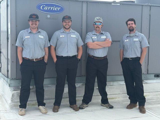 Four HVAC technicians in gray uniforms standing in front of a Carrier unit outdoors.