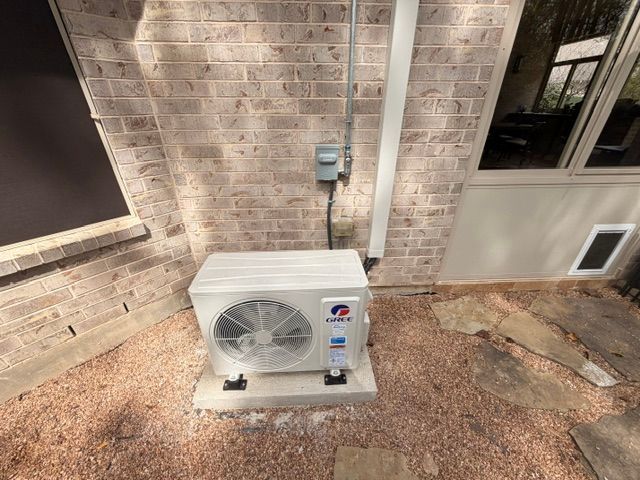 Outdoor HVAC unit beside a brick house wall on a gravel pad with electrical conduit above it