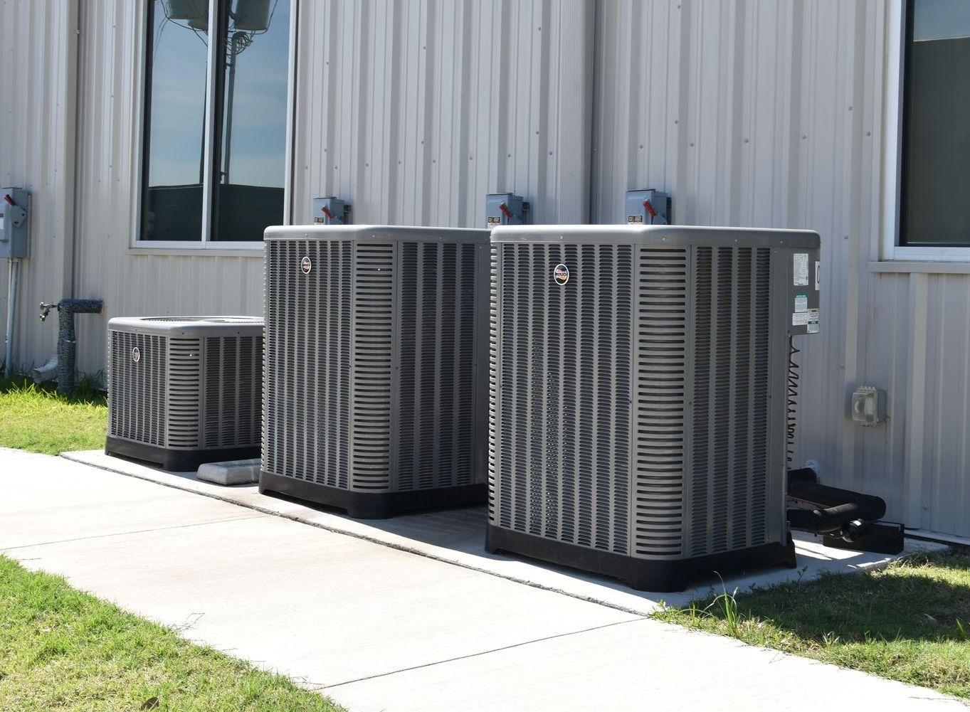 Three air conditioning units outside a building with windows, on a concrete path and grass.