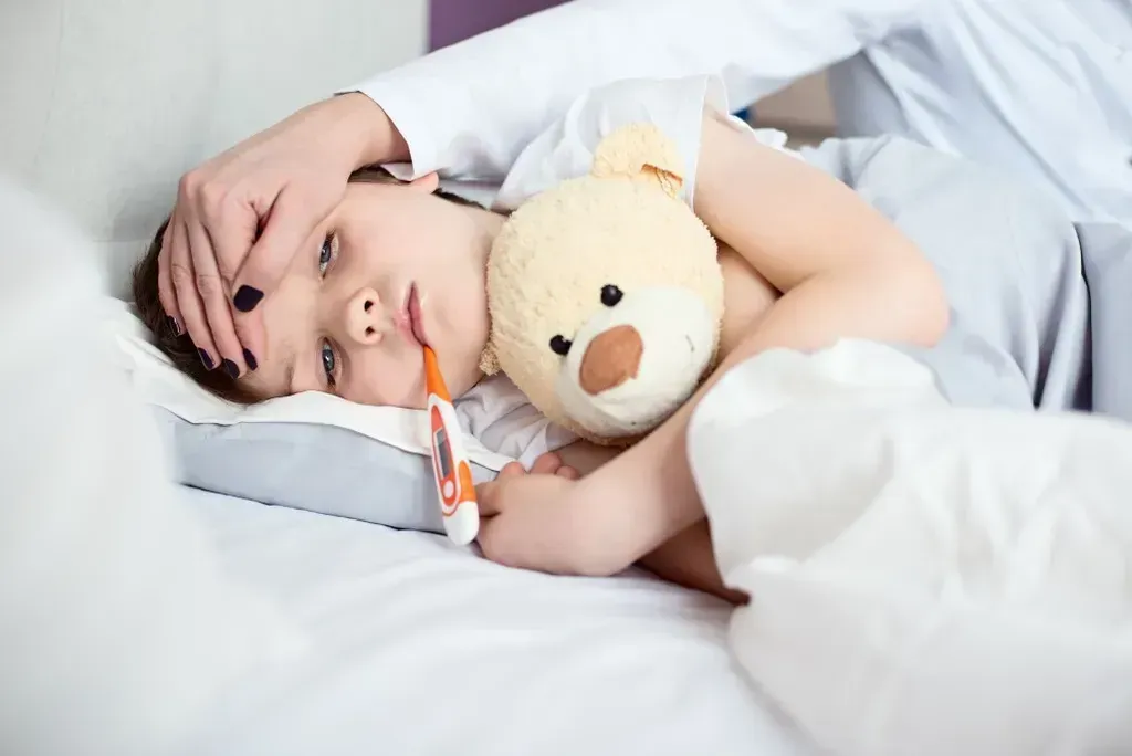 Child laying in bed with a teddy bear and a thermometer in his mouth.