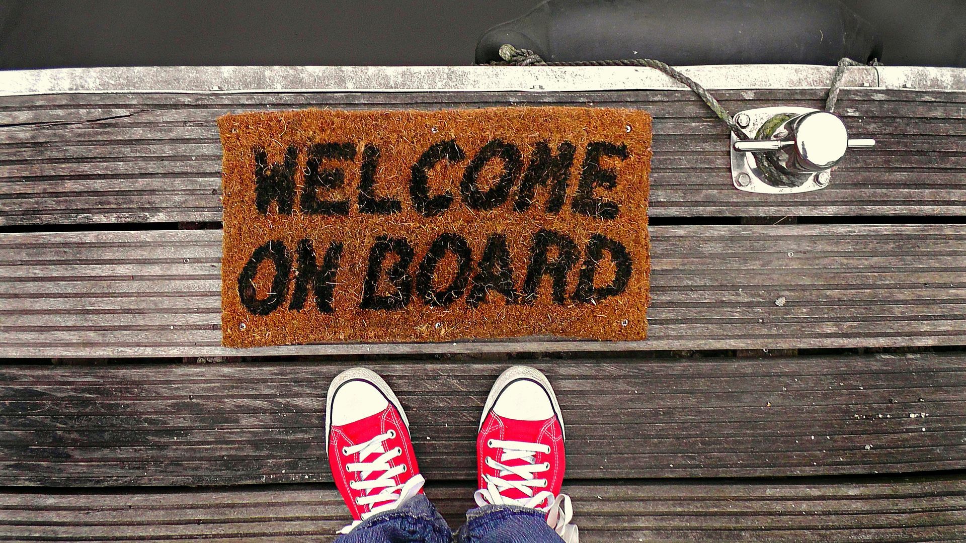 A person is standing on a wooden dock next to a welcome on board doormat.