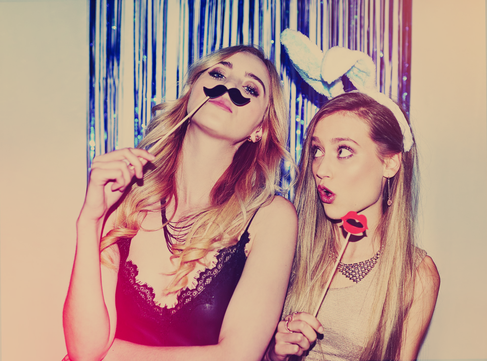 Two women posing with party props: one with a mustache, the other with bunny ears and lips, in front of a blue backdrop.