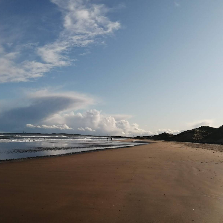 Beach scene under a partly cloudy sky; a person walks along the wet sand.