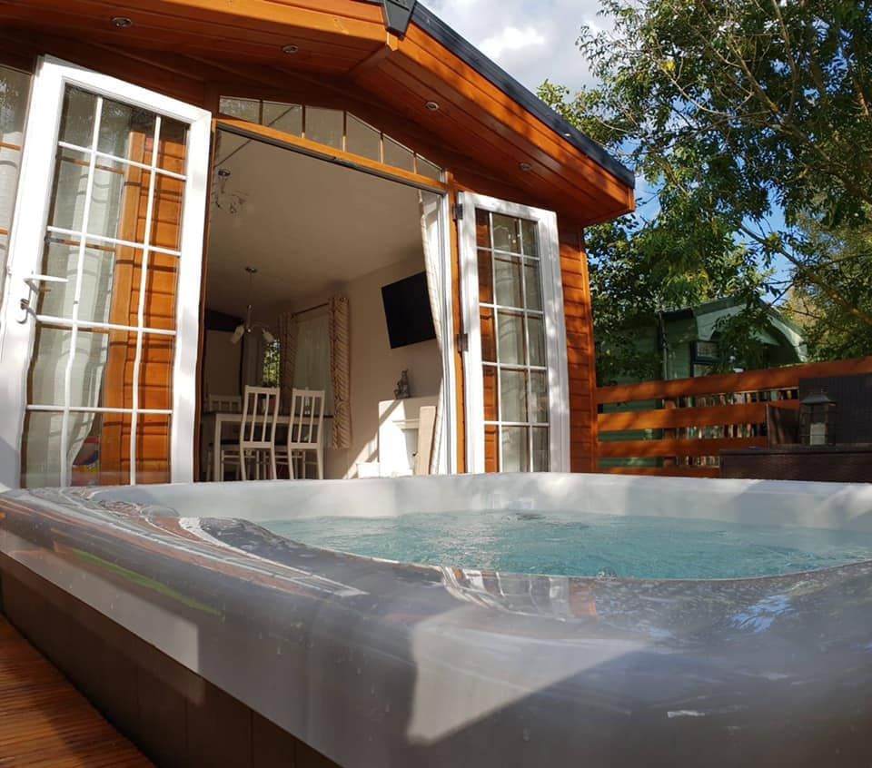 Hot tub on a wooden deck in front of a cabin with open French doors, a table, and a TV inside.