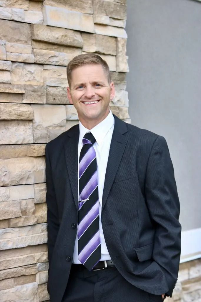 Smiling man in a dark suit and striped tie standing beside a stone wall