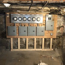 Electrical panels and meters mounted on a wooden board in a basement.