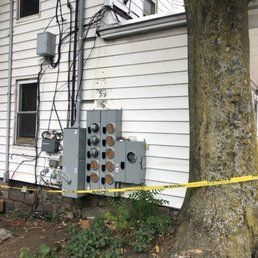 Electrical panel and meters on the side of a white house with yellow caution tape, next to a large tree.
