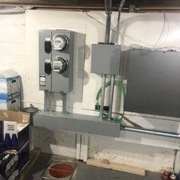 Electrical utility equipment installed on a light-colored wall, consisting of two meters, a box, and conduits.