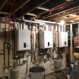Three white tankless water heaters installed on a basement wall with associated plumbing and a stepladder.