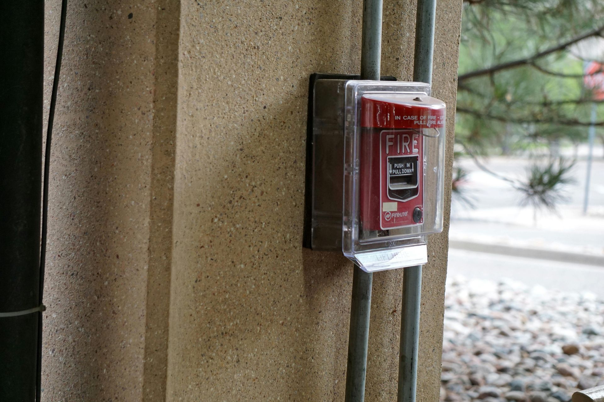 Red fire alarm pull station on a beige concrete pillar, protected by a clear cover. Two gray conduits run vertically.