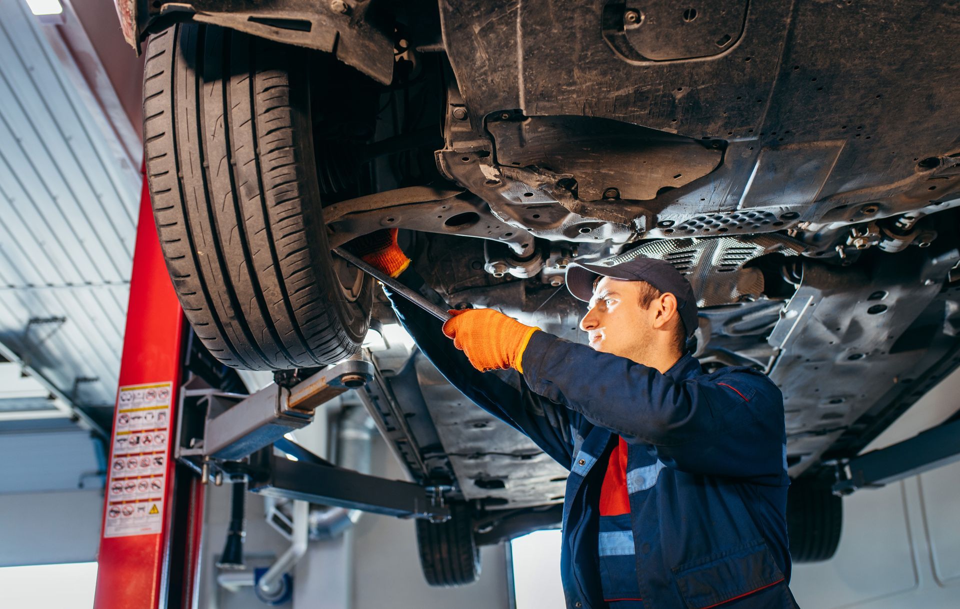 Mechanic under a car on a lift, wearing gloves and overalls, working with tools in a garage.