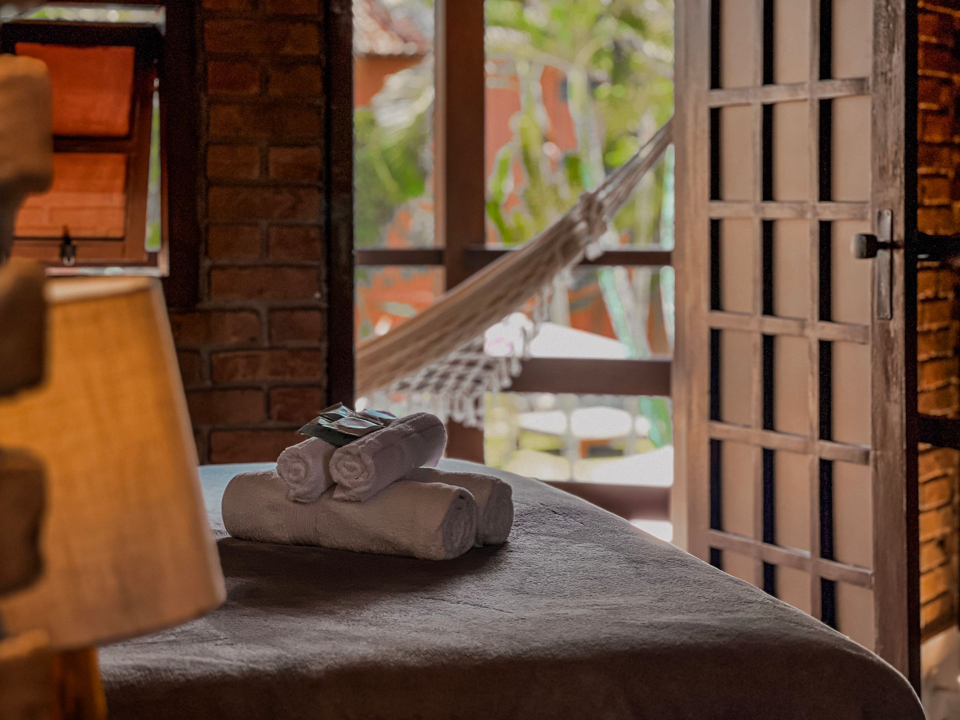 Mesa de massagem com toalhas enroladas, porta aberta para uma varanda com rede. Quarto rústico com parede de tijolos.