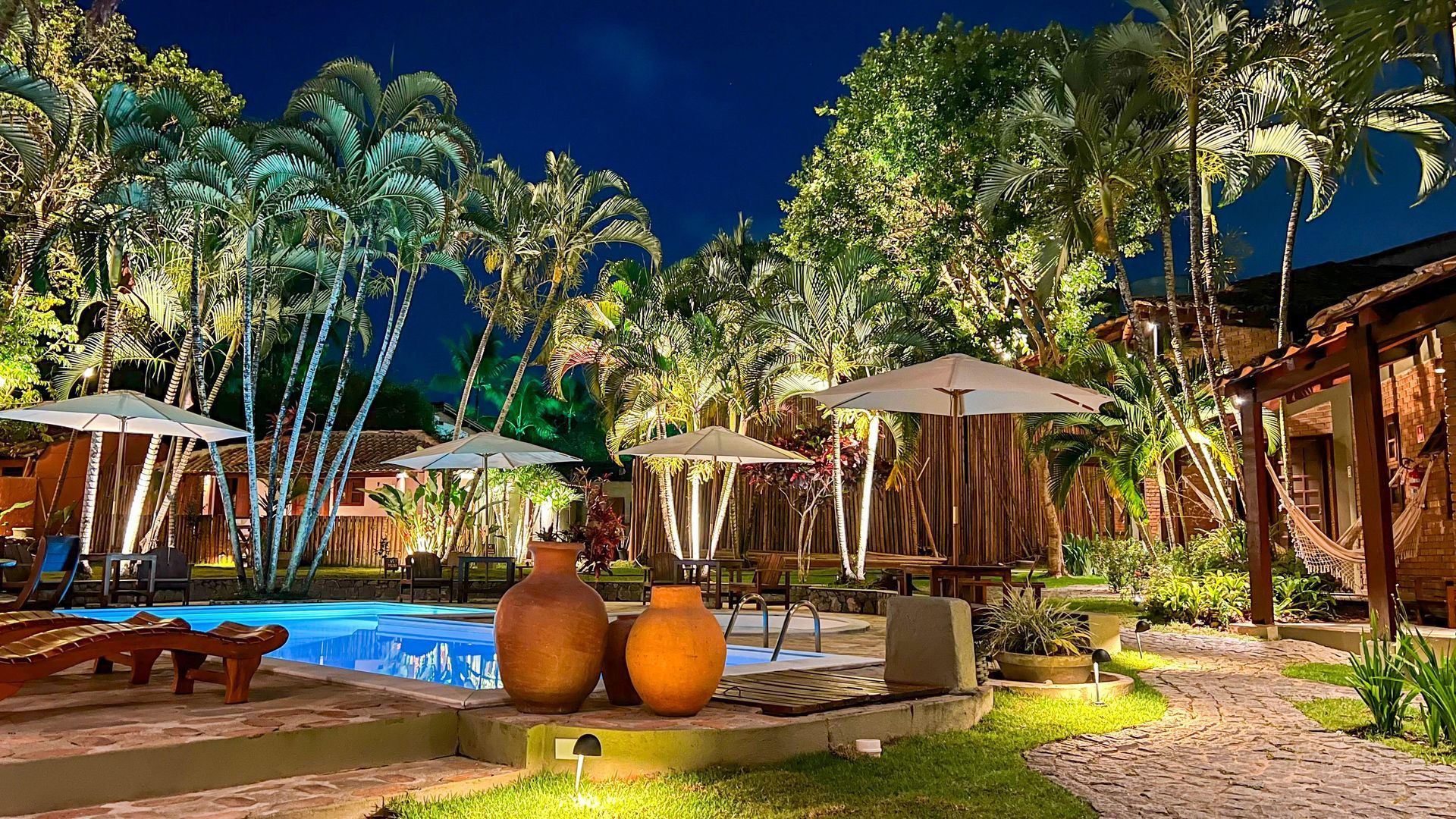Cena noturna à beira da piscina com palmeiras iluminadas, mesas sob guarda-sóis e peças de cerâmica decorativas.
