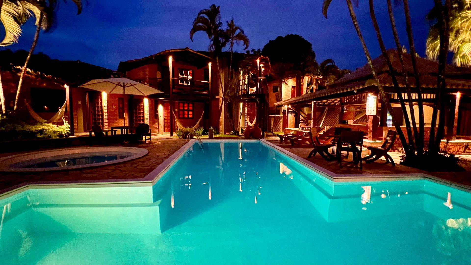 Vista noturna de um resort tropical com piscina iluminada, área de estar ao ar livre e edifícios de vários andares.