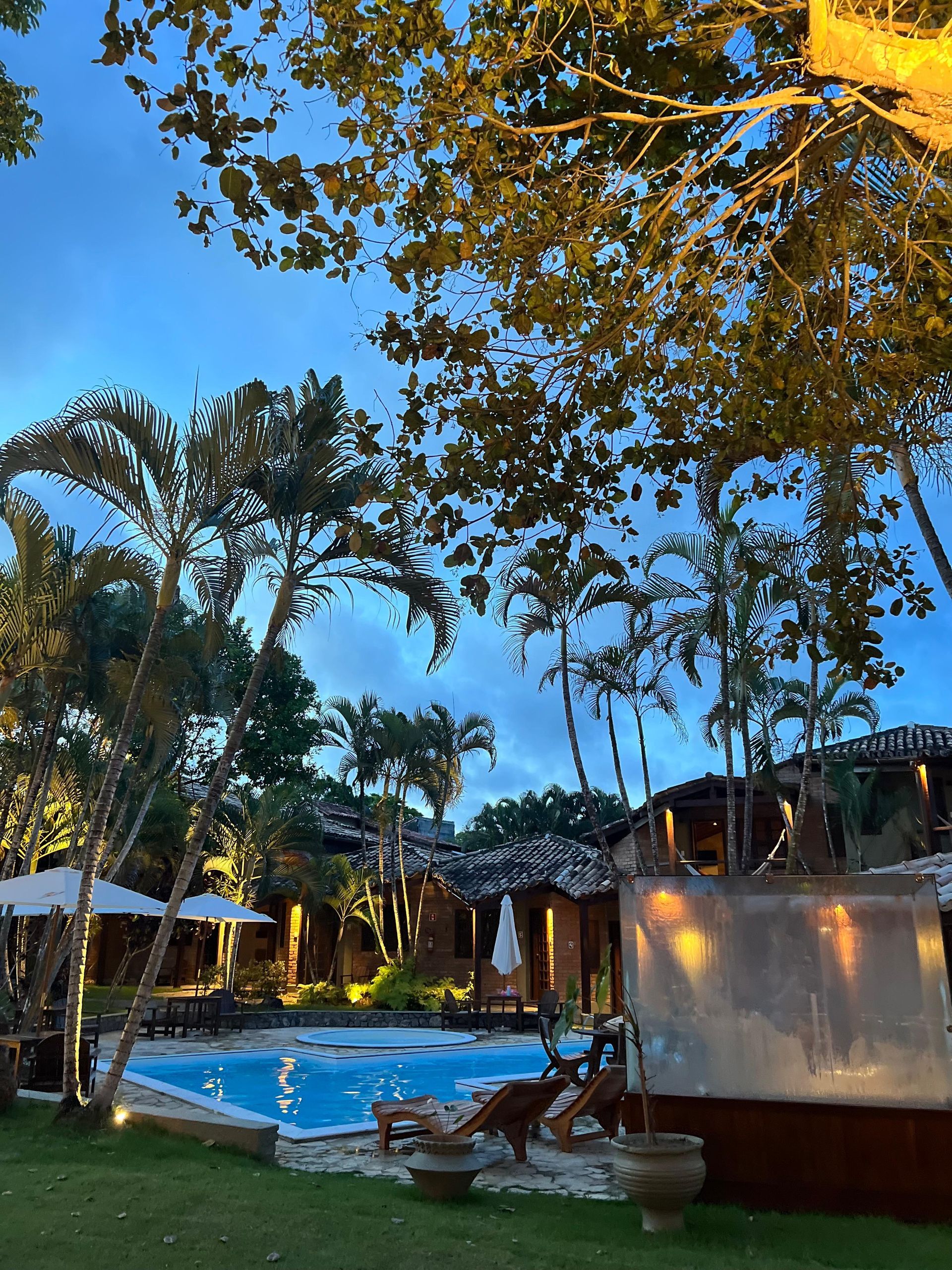 Piscina ao entardecer, rodeada por vegetação exuberante e edifícios iluminados.
