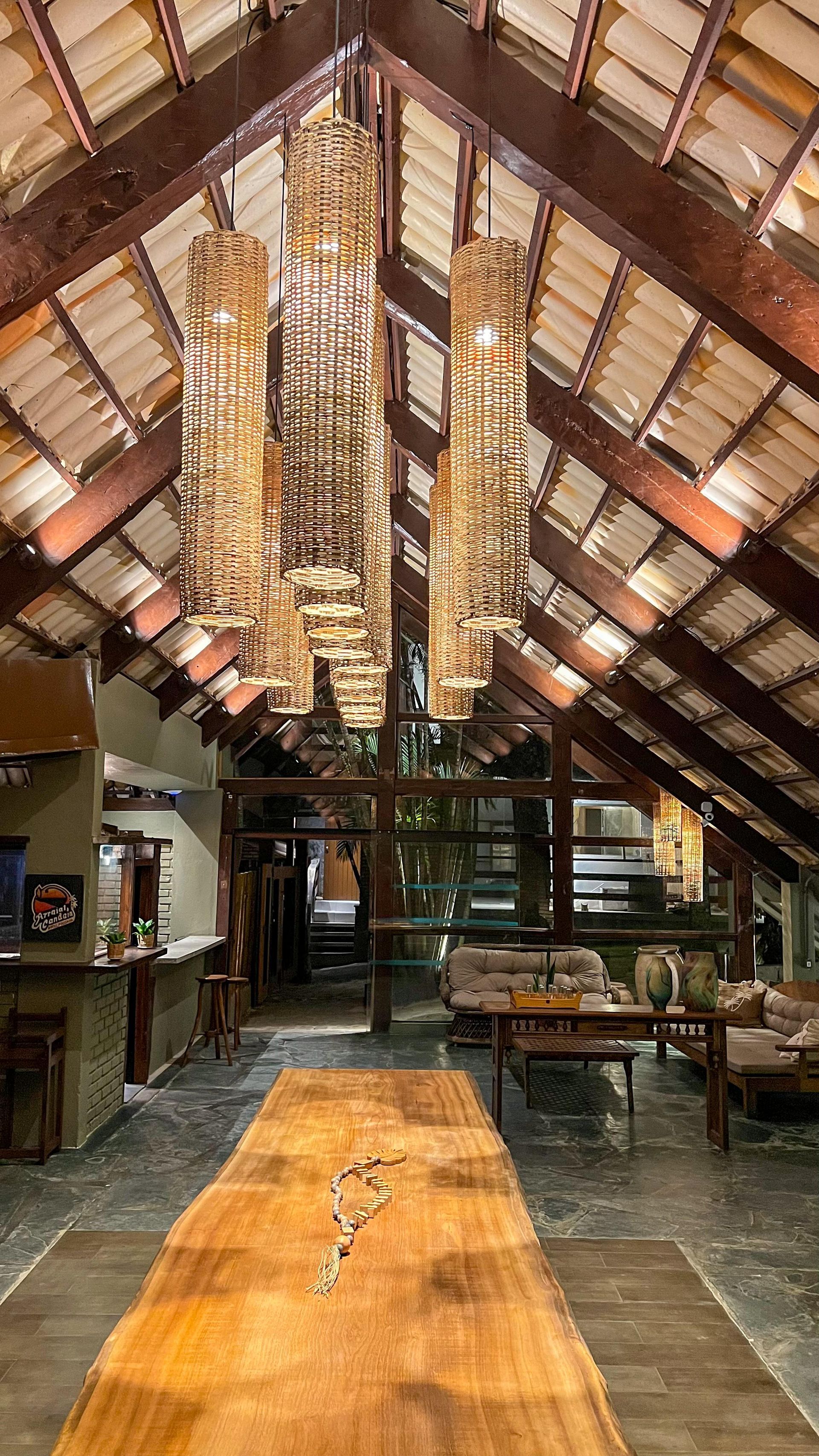 Interior espaçoso com teto alto com vigas aparentes, longa mesa de madeira e luminárias cilíndricas pendentes.