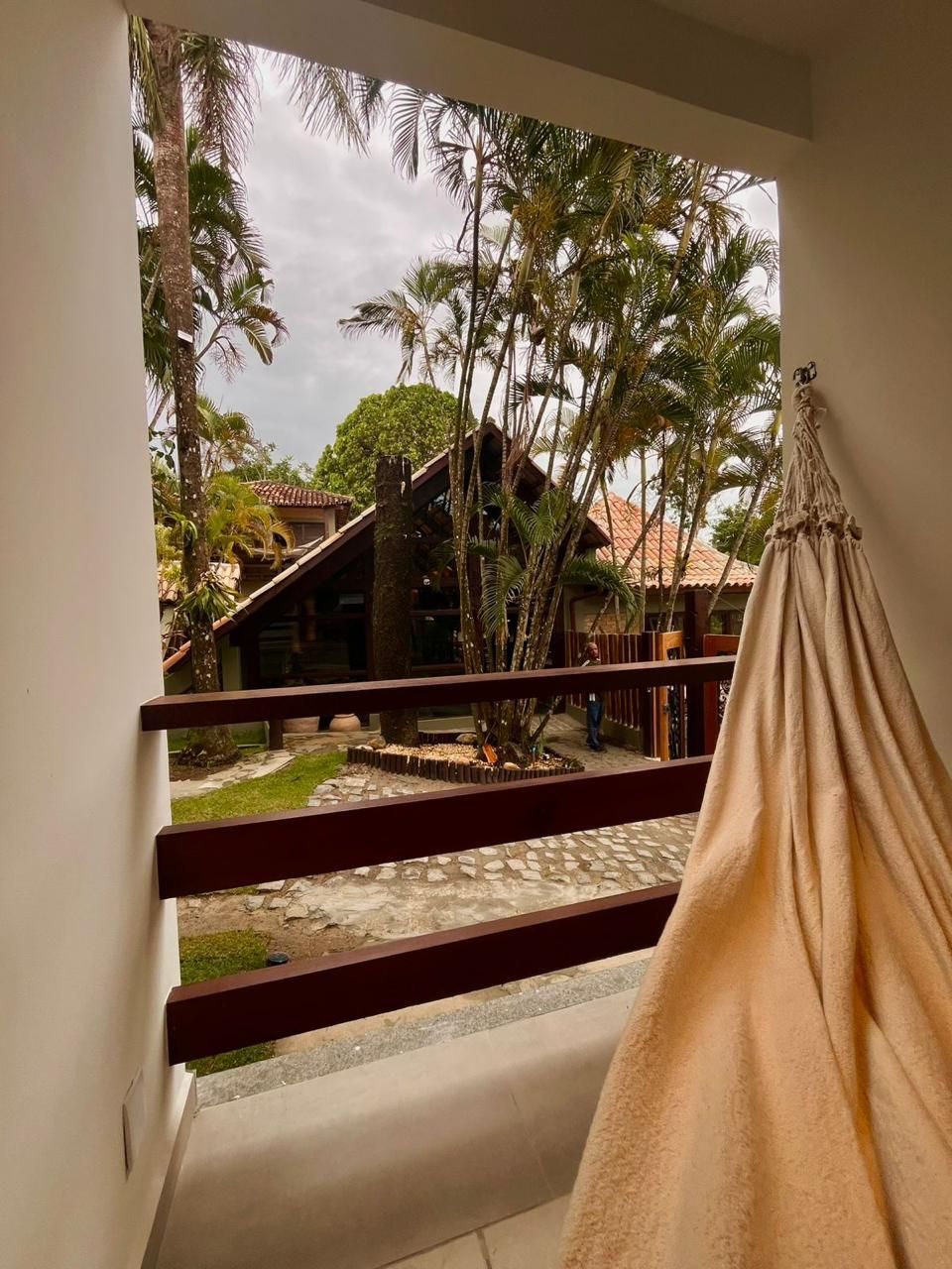 Vista da varanda: uma rede está pendurada perto de um corrimão de madeira. Vista externa de árvores tropicais, estruturas e céu nublado.