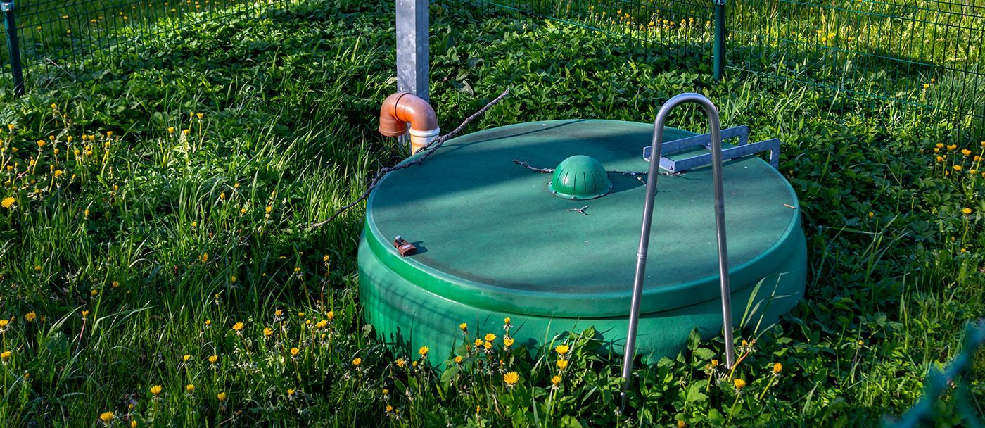 A green septic tank lid sits in a grassy yard, with a metal handle and connected pipes.
