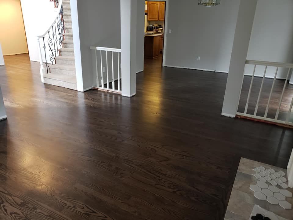 Dark-stained hardwood floors in a home. White columns and stairs with a light-filled kitchen visible in the background.