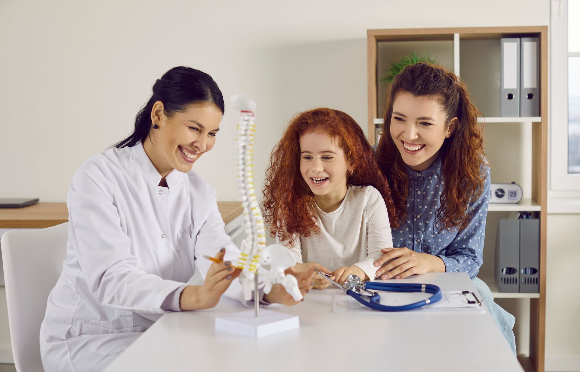 Doctor showing child and parent spinal model, all smiling in an office.