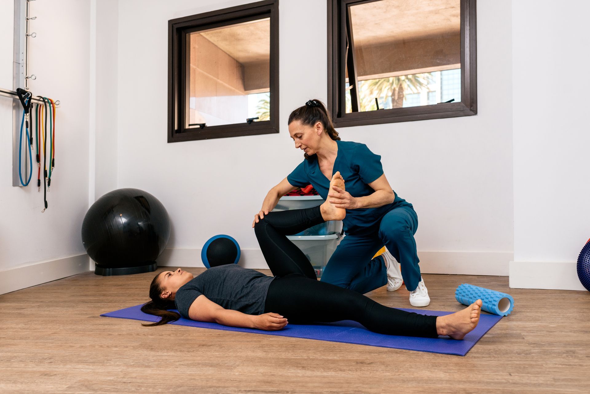 Woman receiving physical therapy from a professional, stretching leg on mat in a clinic.