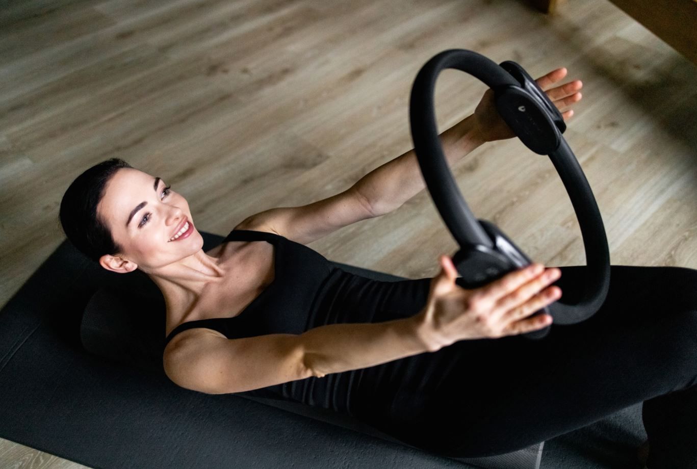 Woman doing Pilates with a ring on a mat, smiling, in a room with light wood flooring.