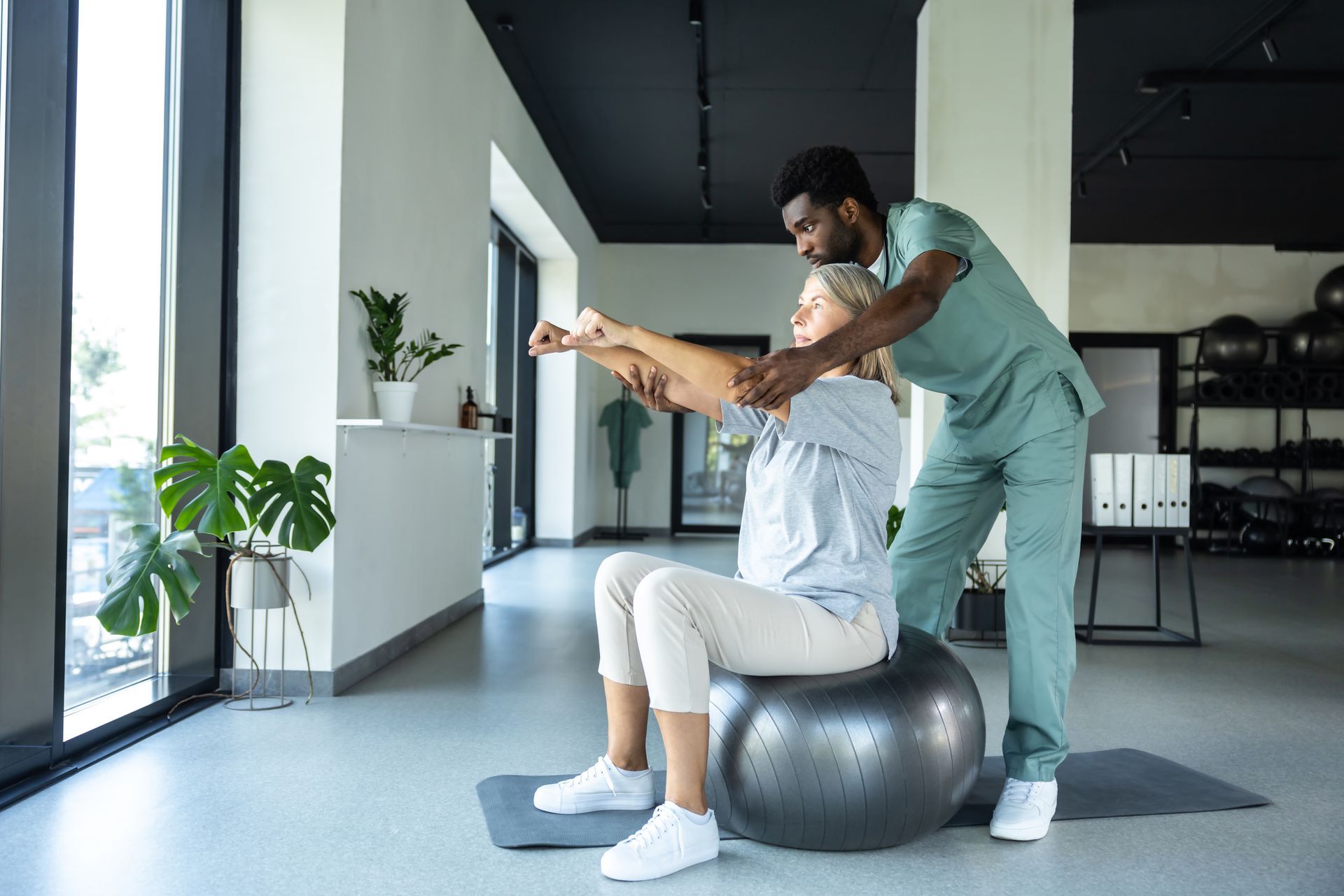 A physical therapist assists a patient with arm exercises while they sit on a stability ball in a gym.