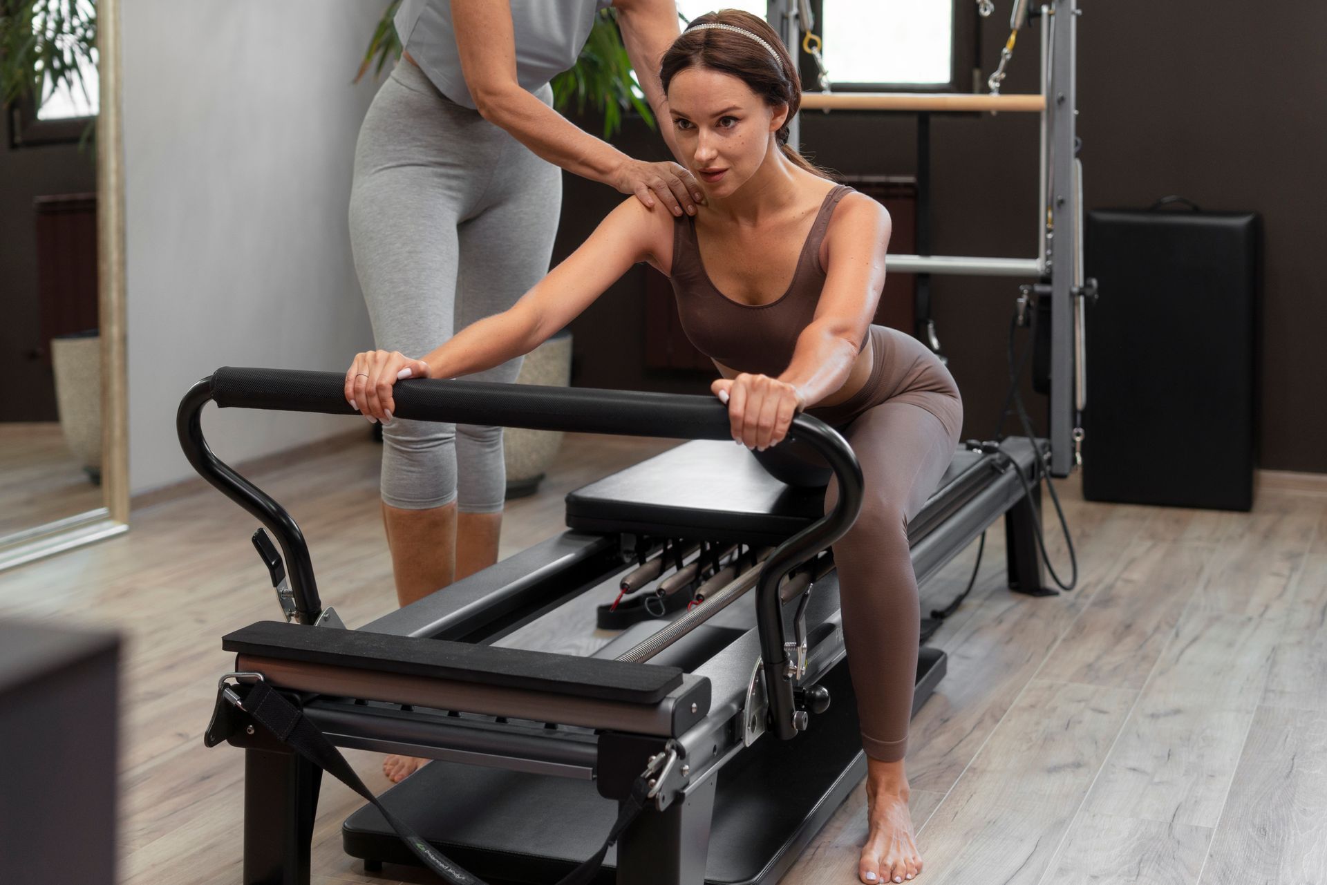 A person performing a Pilates exercise on a reformer machine while a trainer offers support.