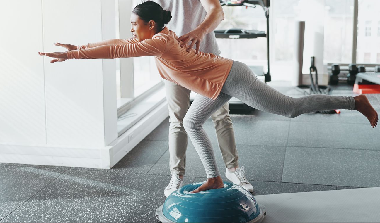 Woman balances on blue exercise ball with trainer's assistance in a gym.