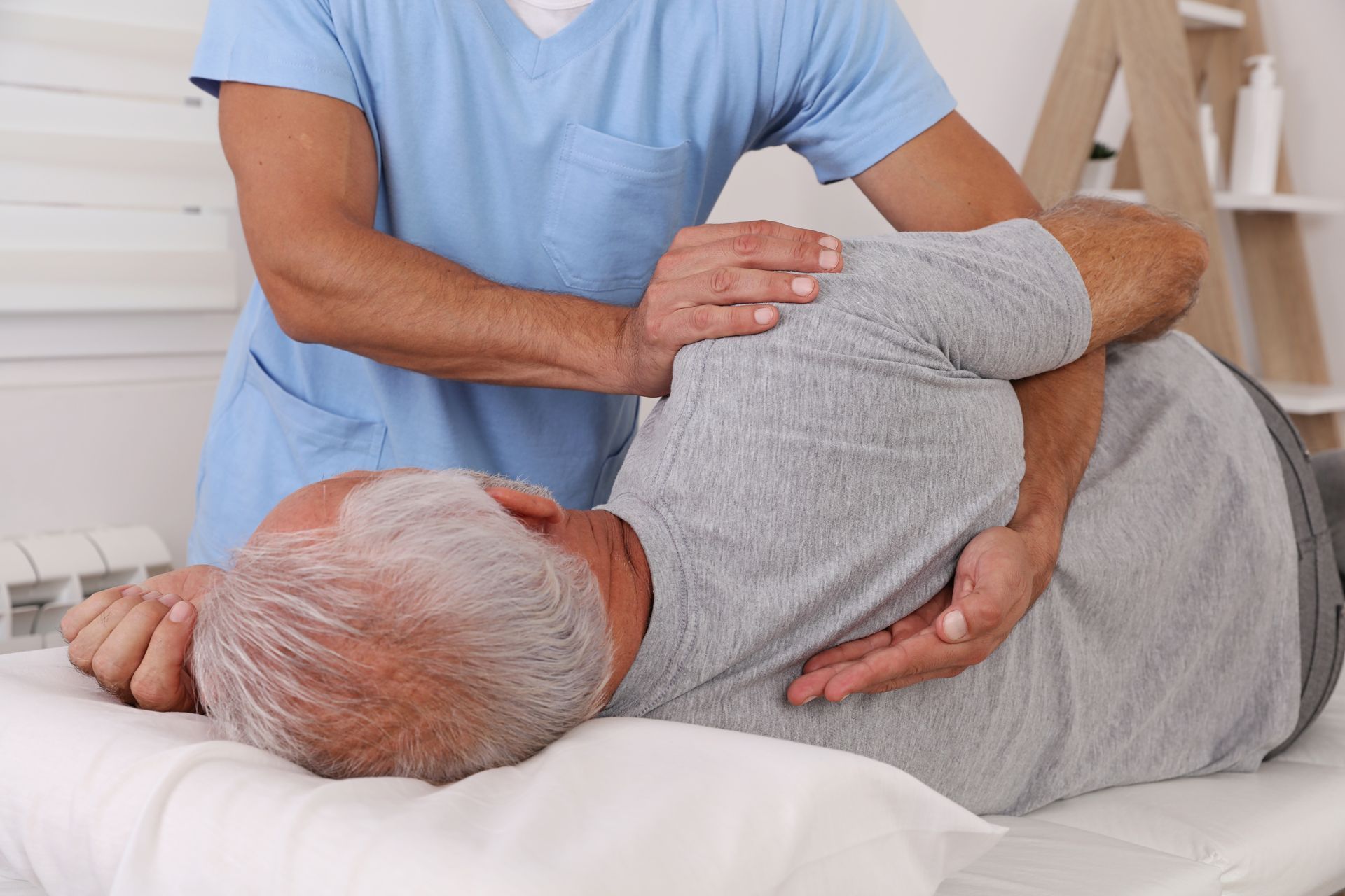 A medical professional in blue scrubs adjusting an older man's back on a massage table.