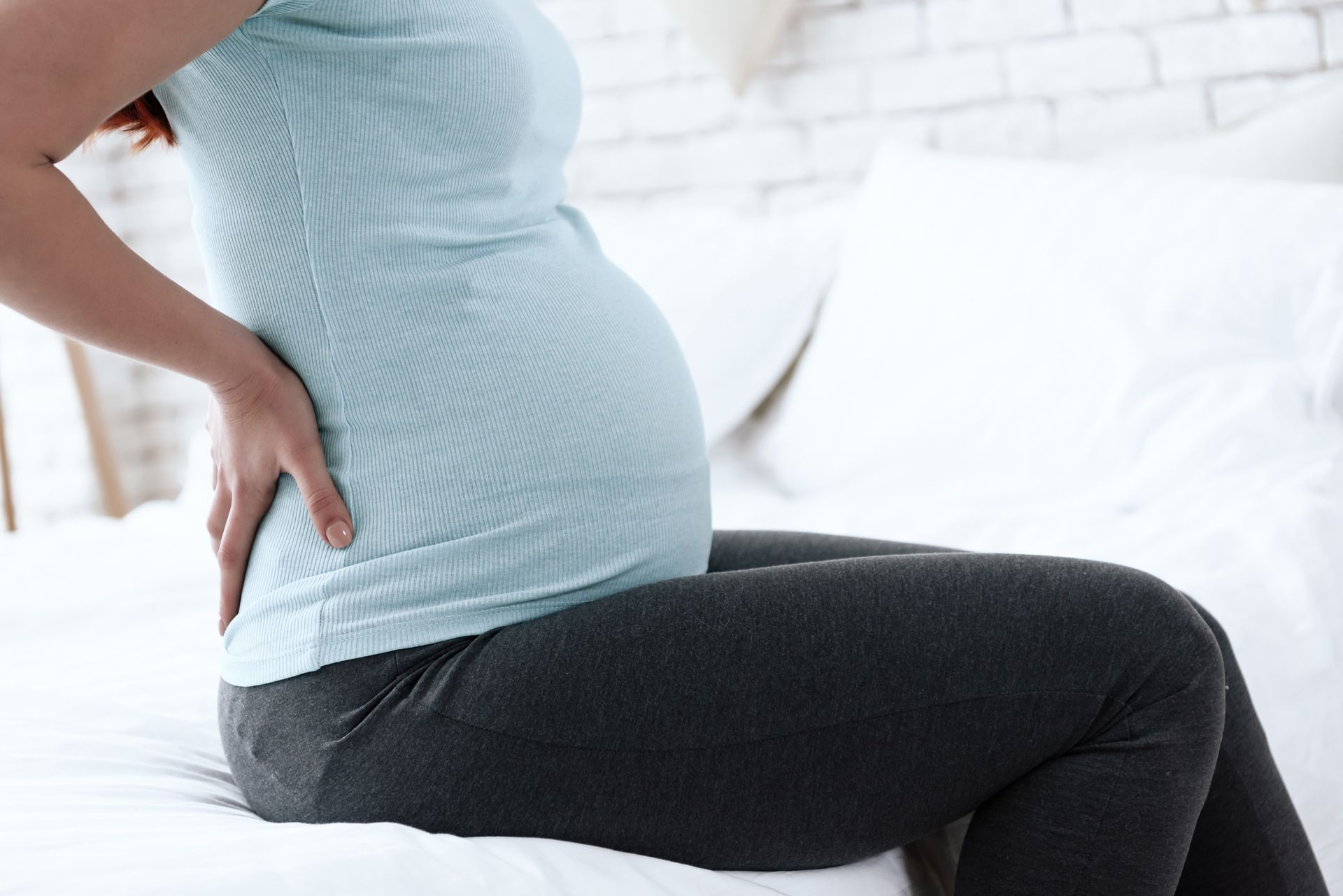 Pregnant woman sitting on a bed, holding her lower back, likely experiencing discomfort.