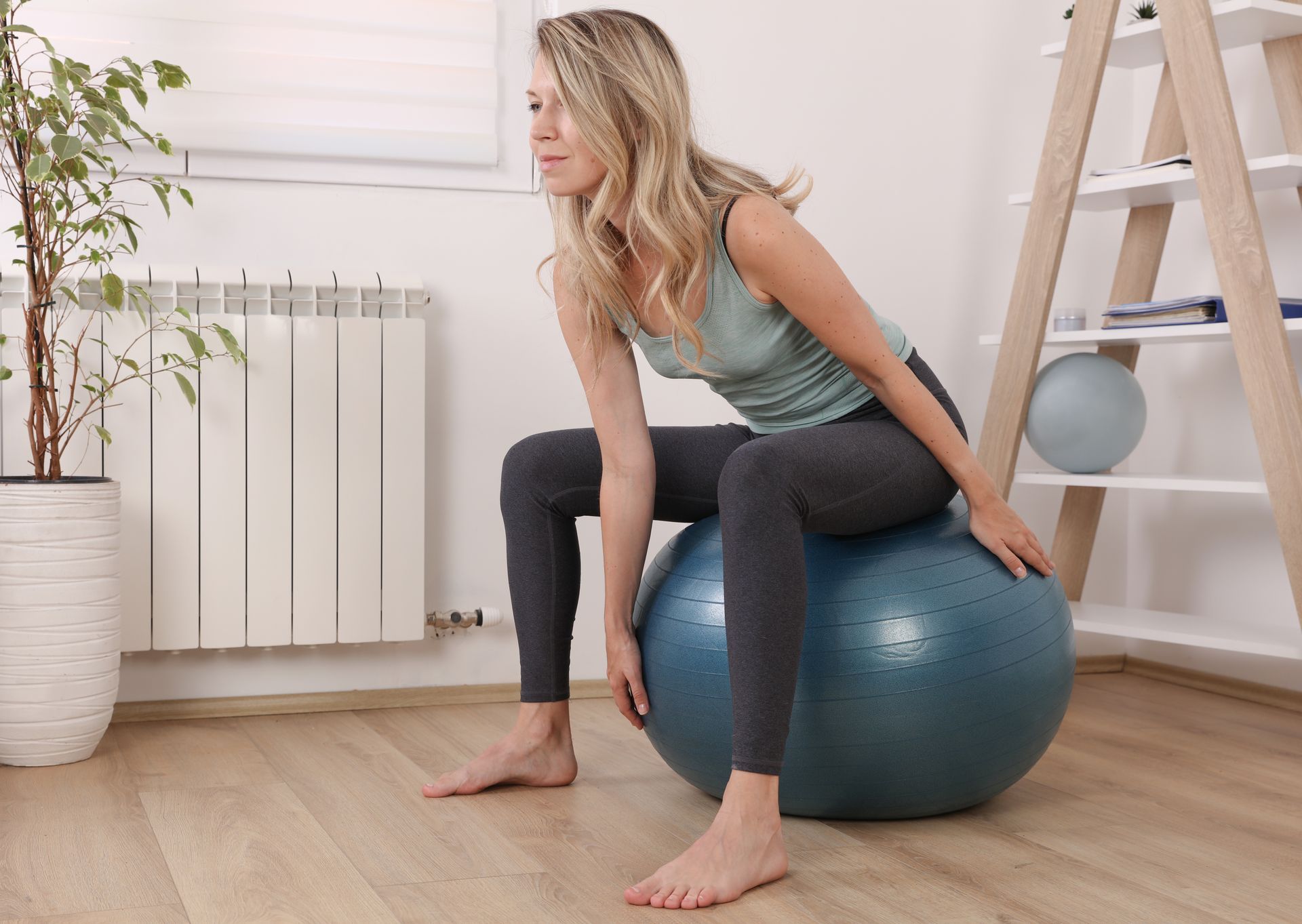 Woman sits on blue exercise ball, arms reaching down. Inside a room, with house plant.
