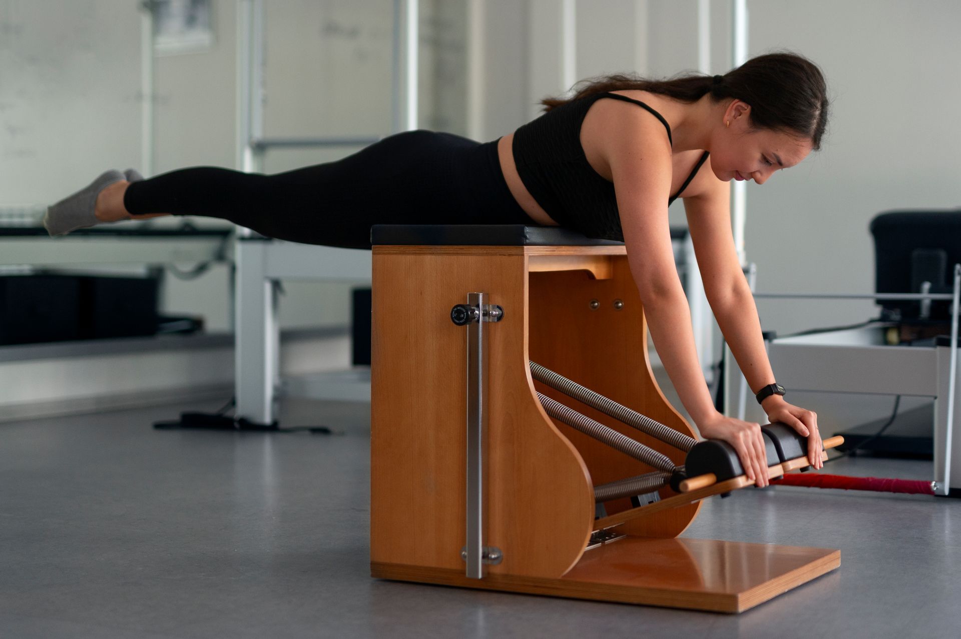 Woman doing pilates exercise on a wooden chair, arms extended, body horizontal, in a studio.