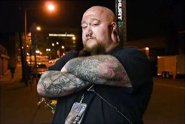 A man with a beard and tattoos is standing on a street with his arms crossed.