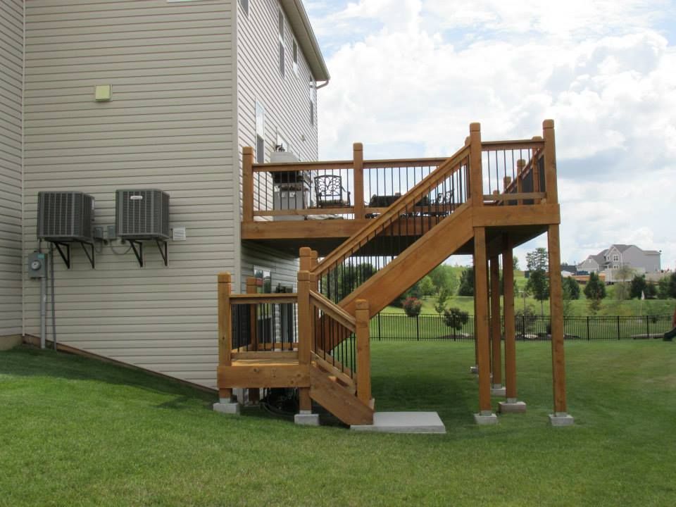 Wooden two-story deck with stairs attached to a beige house, set on green grass under a cloudy sky.