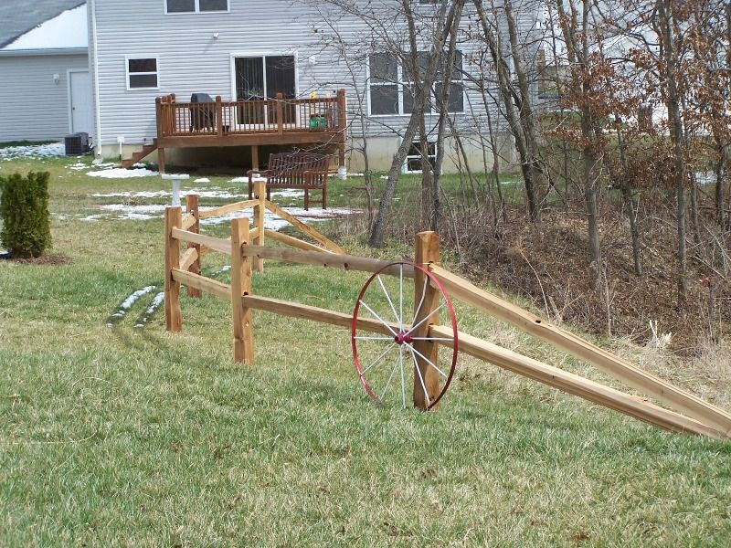 Wooden split-rail fence in backyard with a wagon wheel hung on it, and a house in the background.